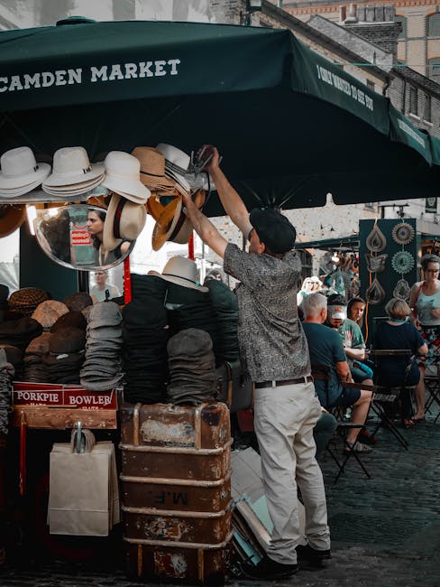 A street vendor at Camden Market arranging hats on display under a large green canopy with white lettering. The hats are of various styles and colors, neatly stacked on tables. The canopy, featuring t