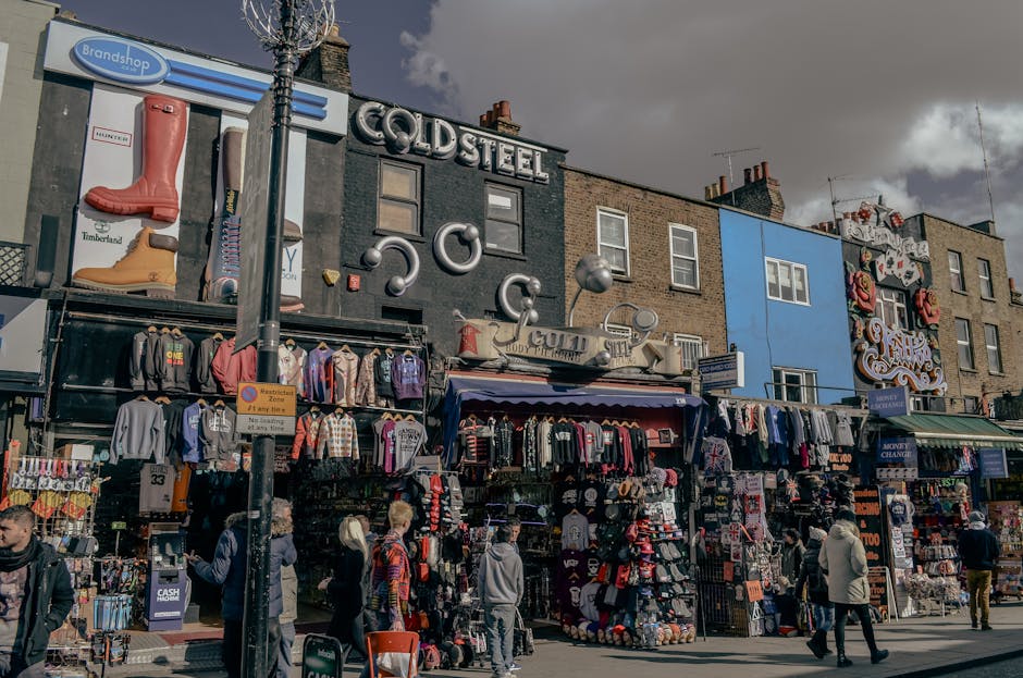 A street vendor at Camden Market arranging hats on display under a large green canopy with white lettering. The hats are of various styles and colors, neatly stacked on tables. The canopy, featuring t