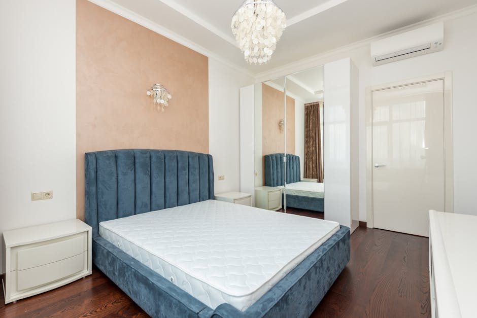 A modern, minimalist bedroom with white painted walls and a grey upholstered bed frame topped with a clean, white mattress. The room features a built-in wardrobe with beige doors and dark wood accents